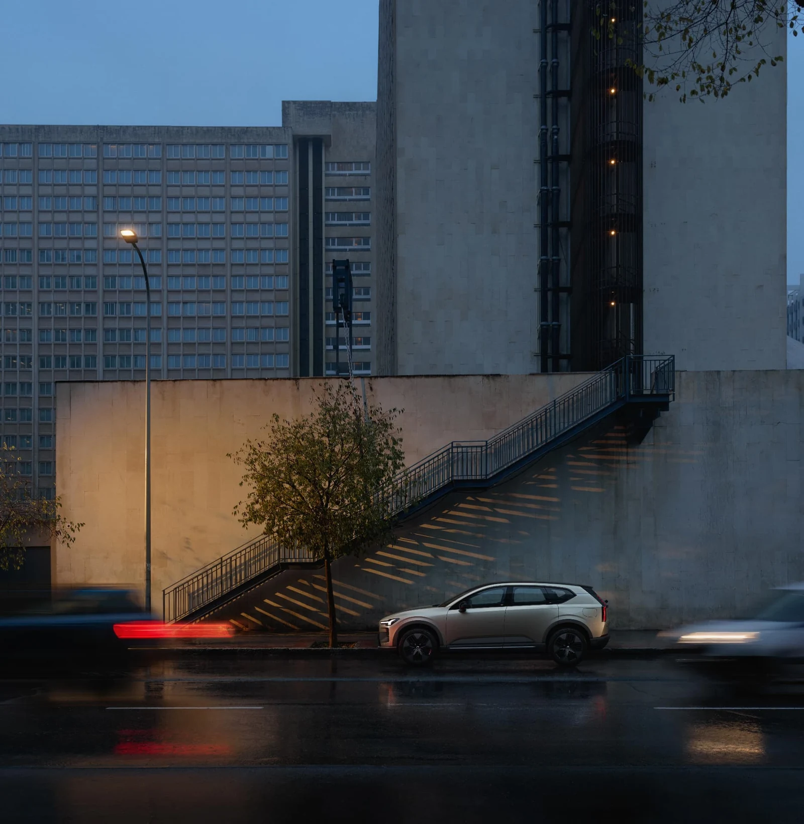 A Volvo parked on a wet city street at dusk, traffic passing in long exposure