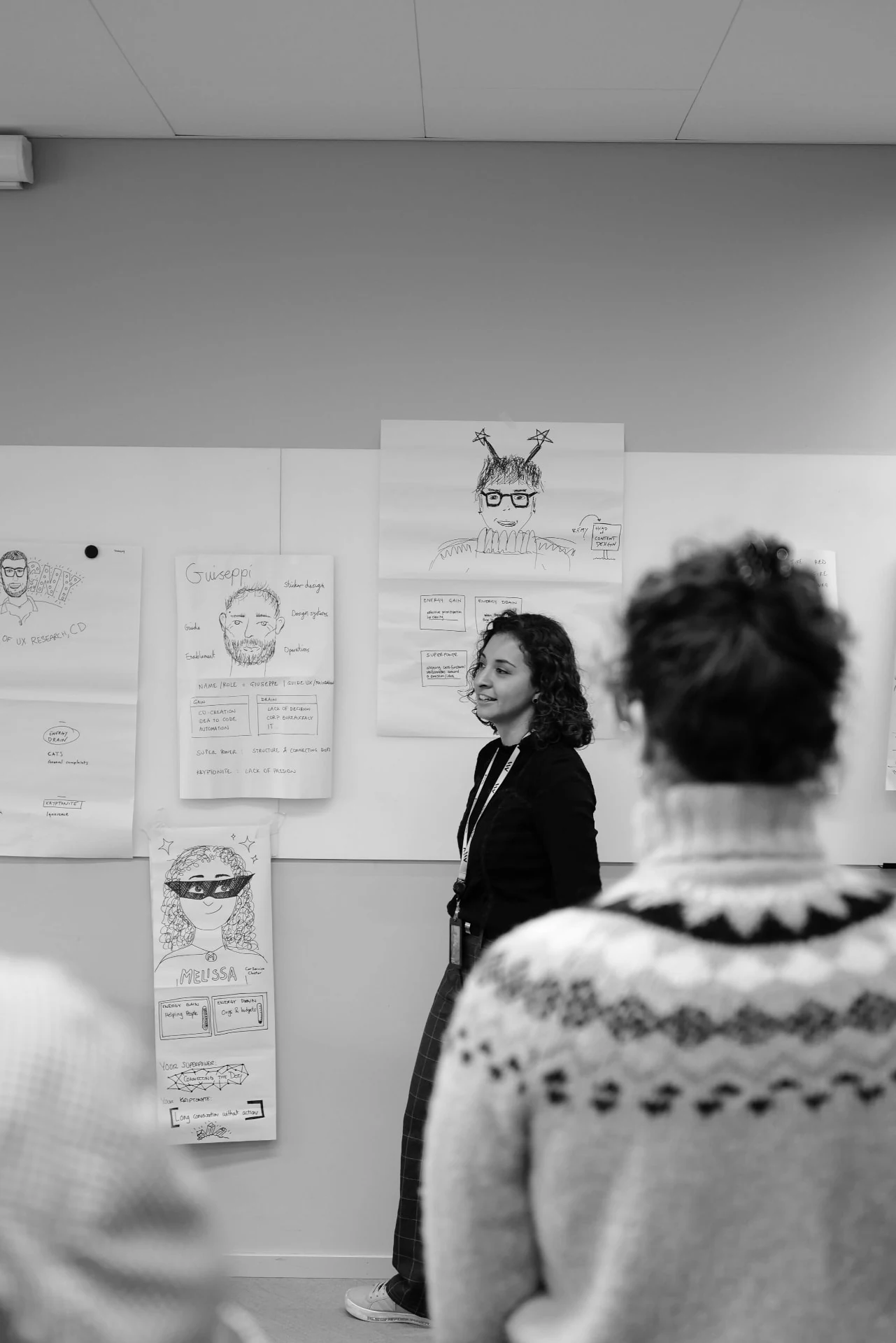 A colleague speaking beside hand-drawn character sheets pinned to the wall