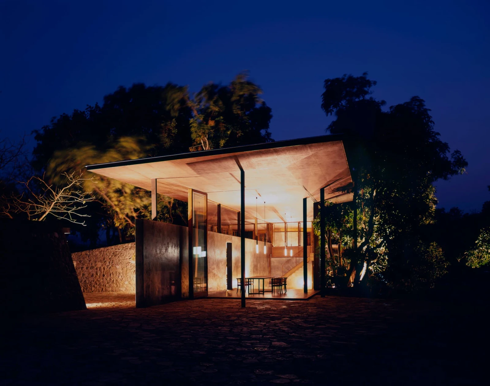 Open pavilion lit from within at dusk, pendant lights glowing under a wide concrete roof